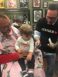 A baby in a man's lap receives a haircut from a barber